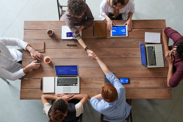 Reunión de equipo de negocios vista desde arriba, con varias personas trabajando en laptops alrededor de una mesa de madera y dos de ellas dándose la mano al centro como señal de acuerdo.