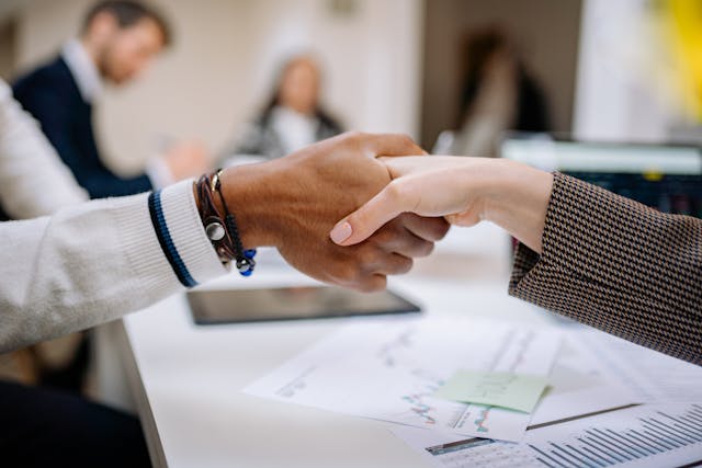Dos personas sellando un acuerdo con un apretón de manos en una reunión de negocios, sobre una mesa con gráficos financieros y documentos de estrategia.