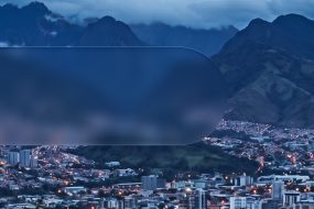 Vista nocturna de una ciudad latinoamericana rodeada de montañas, con edificios iluminados y paisaje montañoso de fondo.