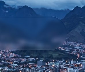 Vista nocturna de una ciudad latinoamericana rodeada de montañas, con edificios iluminados y paisaje montañoso de fondo.