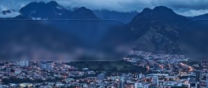 Vista nocturna de una ciudad latinoamericana rodeada de montañas, con edificios iluminados y paisaje montañoso de fondo.