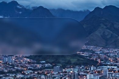 Vista nocturna de una ciudad latinoamericana rodeada de montañas, con edificios iluminados y paisaje montañoso de fondo.